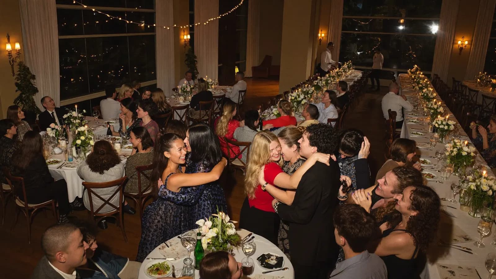 A class reunion in a candlelit banquet hall: old classmates greeting each other with joyful hugs between round tables